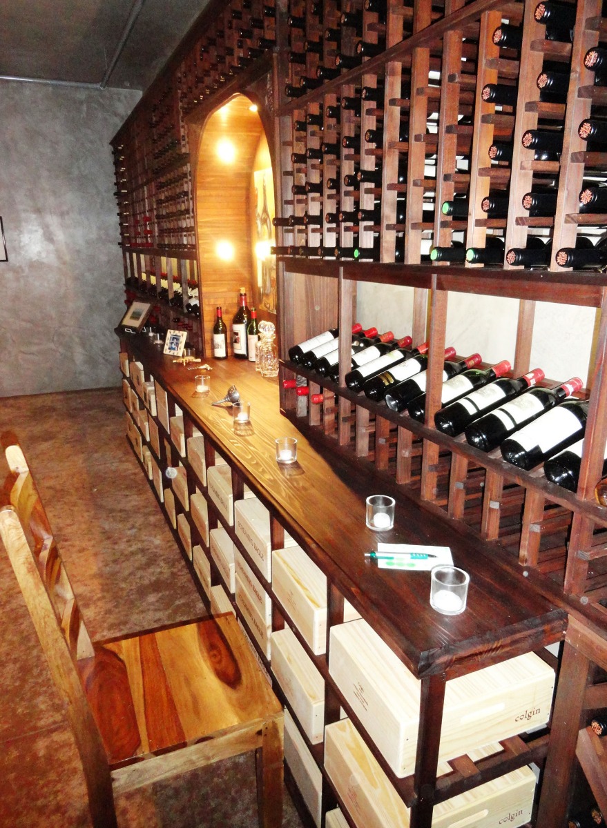Long tasting countertop with wine case storage and diamond bins below, topped by a high reveal angled display and library wall of single deep wine bottles